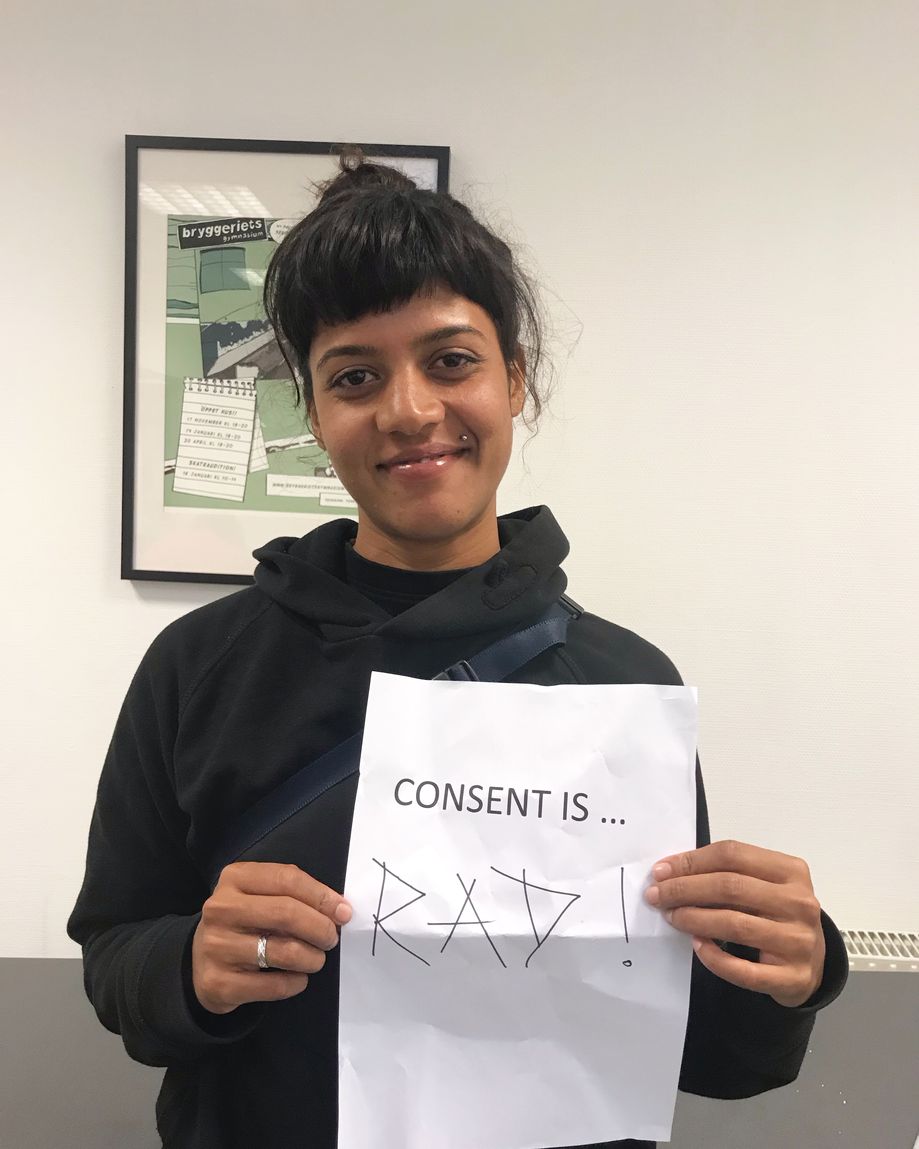 Atita, from India, is dressed in black and indoors holding up a piece of paper saying Consent is Rad.