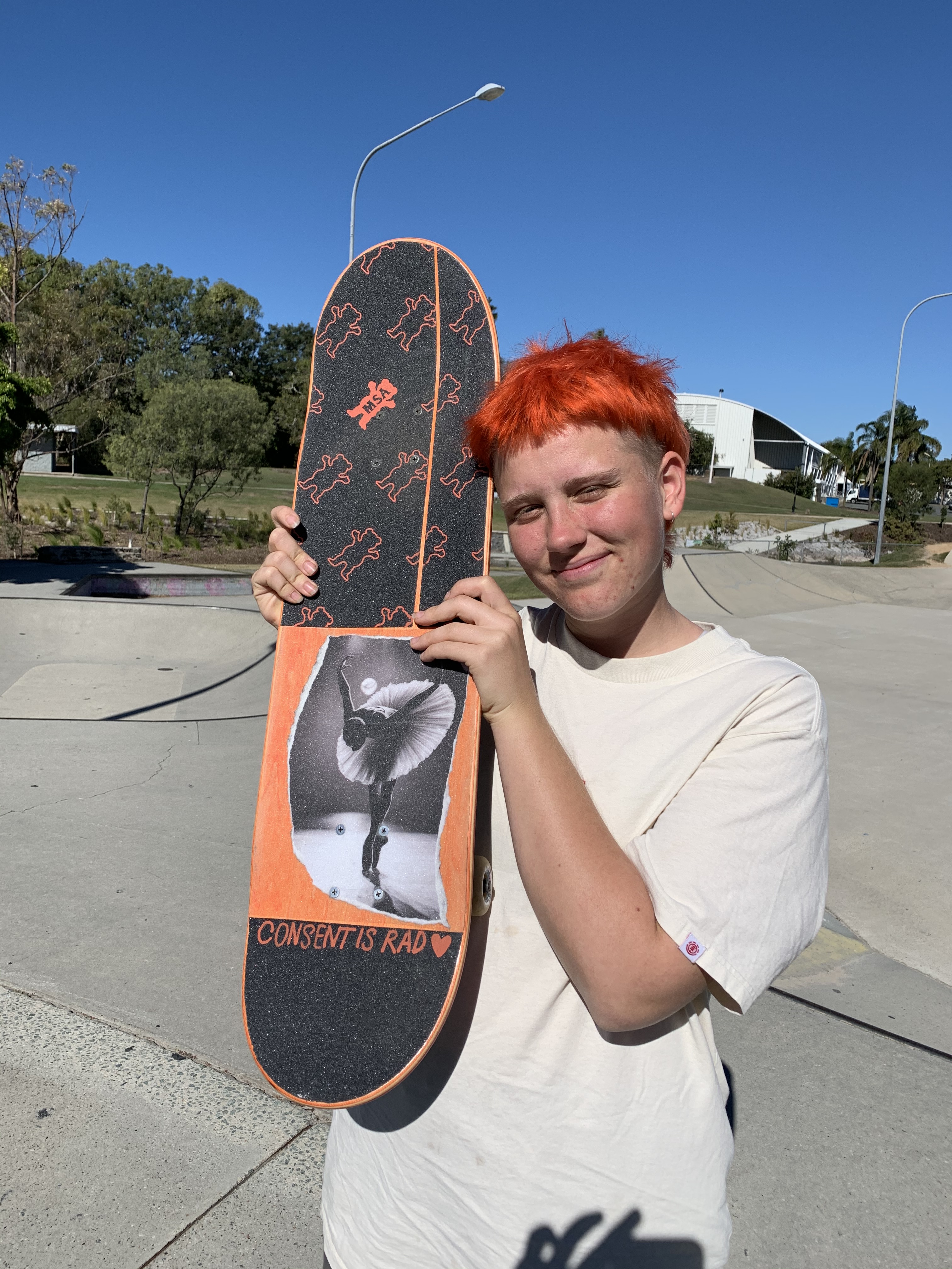 Millie is outdoor at a skatepark with a skate deck saying consent is rad in the sun. Her orange grip tape matches her